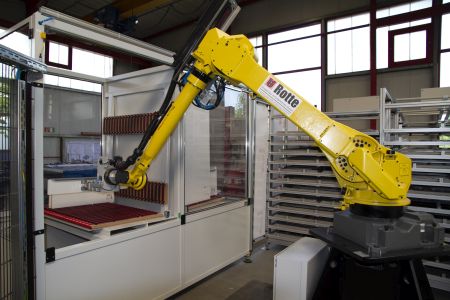 Gelber Industrieroboterarm der Marke Rotte in einer Fertigungshalle, der ein Bauteil greift, umgeben von Maschinen und Metallgestellen.