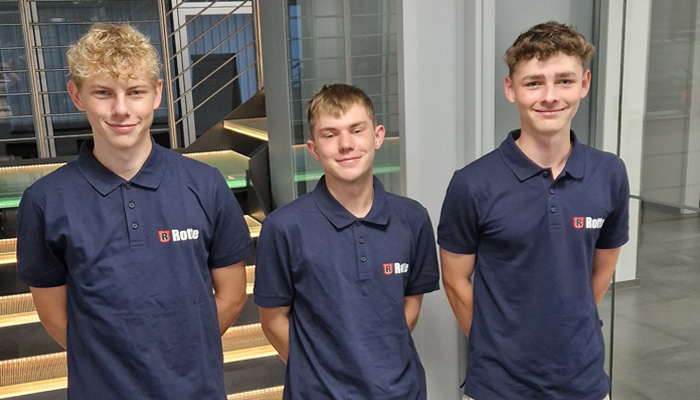 Drei junge Männer in blauen Poloshirts mit dem Logo 'R Rotte' stehen vor einer modernen Treppe aus Glas und Metall, alle lächeln leicht und haben die Hände hinter dem Rücken.