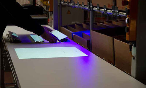 Person legt Papier mit UV-Marker unter eine blaue UV-Lampe auf einem Arbeitstisch in einer Lagerumgebung mit Kartons.
