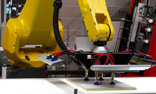 Nahaufnahme eines gelben Industrieroboterarms, der eine flache Platte mit Saugern greift, in einer Werkstatt oder Produktionsumgebung
