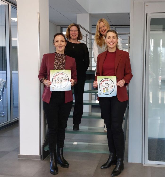 Vier Frauen auf einer Treppe, zwei vorne mit roten Blazern und Haltepunkten-Zertifikaten in den Händen.