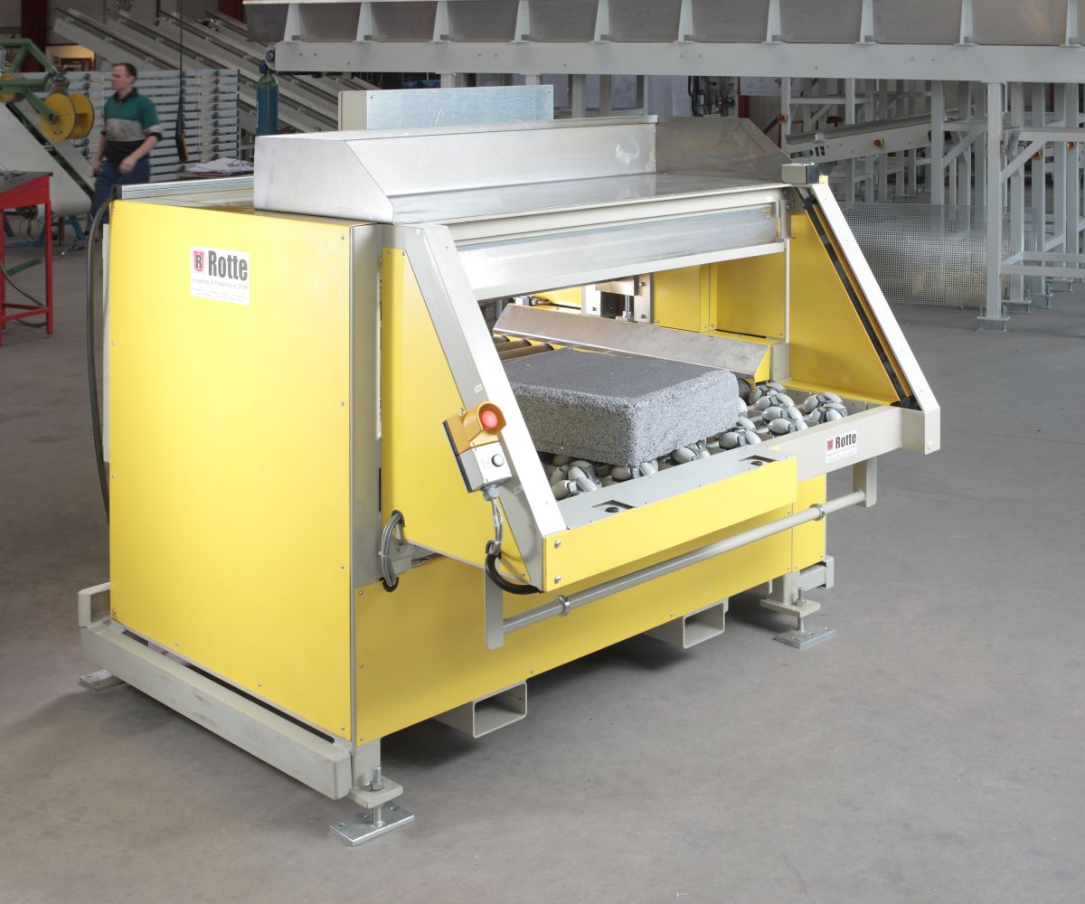 gelbe Industriemaschine mit Förderband und grauem Schaumstoffblock in Werkhalle, im Hintergrund Person und Industrieanlagen