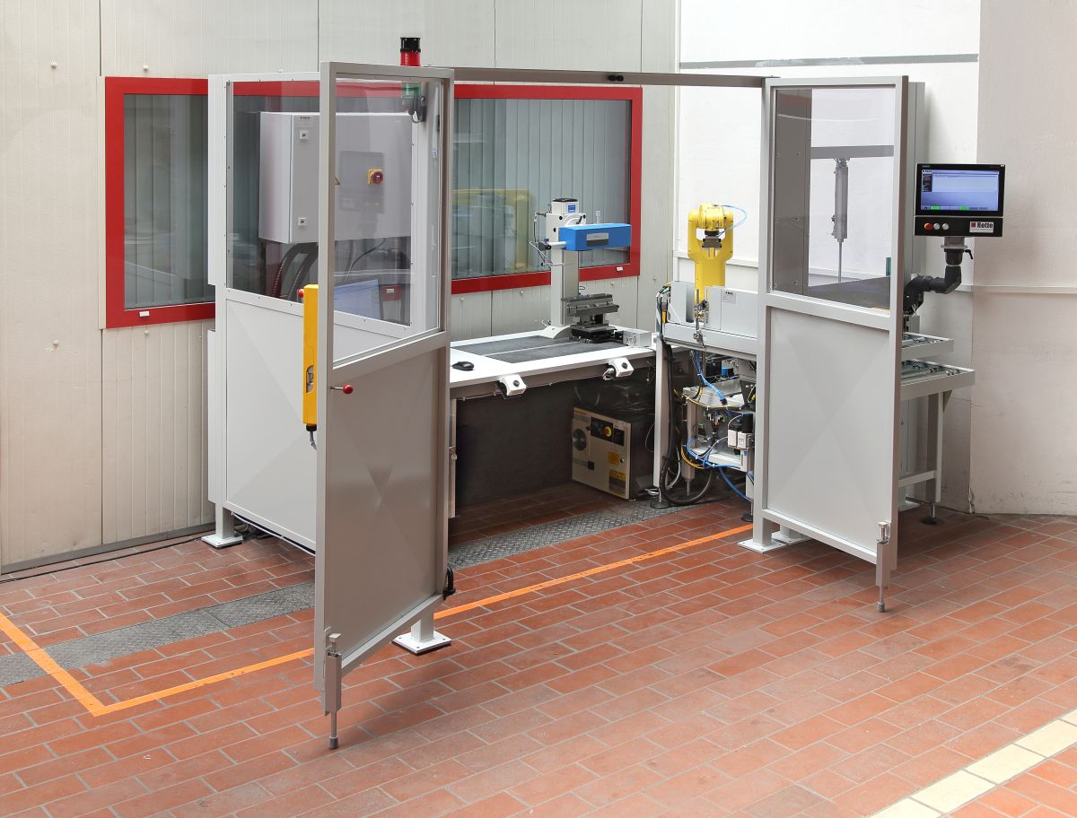 Industrielle Mess- oder Prüfstation mit offener Schutzkabine, Monitor, gelbem Roboterarm und Messgerät auf schwarzem Tisch im Raum mit rotem Fensterrahmen.