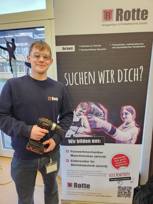 Junger Mann mit Schutzbrille und Akku-Bohrmaschine steht neben einem Werbebanner der Firma Rotte für Ausbildungsberufe in Feinwerkmechanik, Maschinenbau und Elektronik.