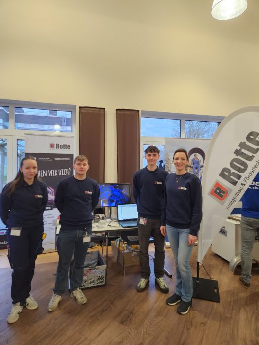 Vier junge Erwachsene in blauen Pullovern mit Firmenlogo stehen vor einem Messestand mit Bannern und Monitoren in einem Innenraum.