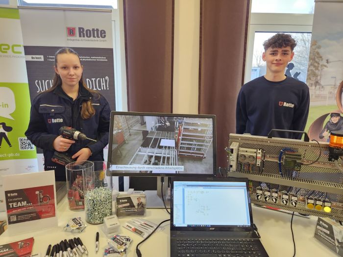 Zwei Jugendliche in blauer Arbeitskleidung stehen an einem Messestand mit Computer, Werkzeug und Infomaterialien einer Firma namens Rotte.