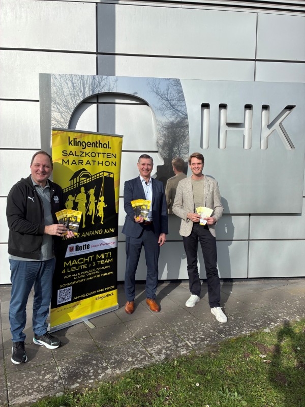 Drei Männer vor einem IHK-Gebäude mit einem Banner zum Salzkotten Marathon, sie halten Flyer in der Hand.