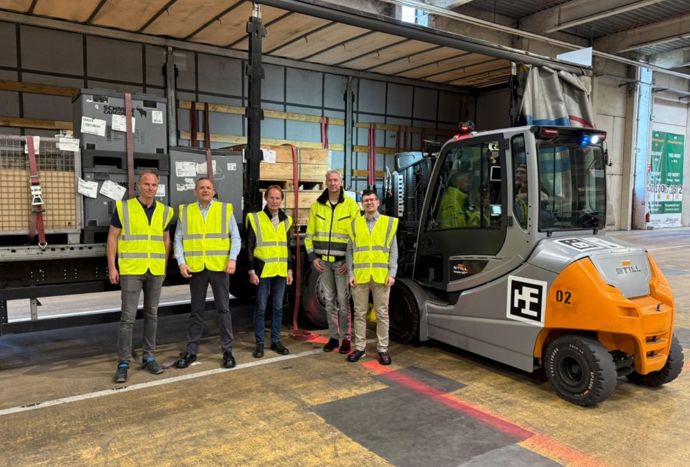 Fünf Männer mit gelben Sicherheitswesten neben einem Gabelstapler und einem beladenen LKW in einer Lagerhalle.