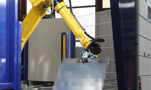 Gelber Industrieroboterarm mit Greifer manipuliert ein unscharf dargestelltes Objekt in einer Industrieumgebung.