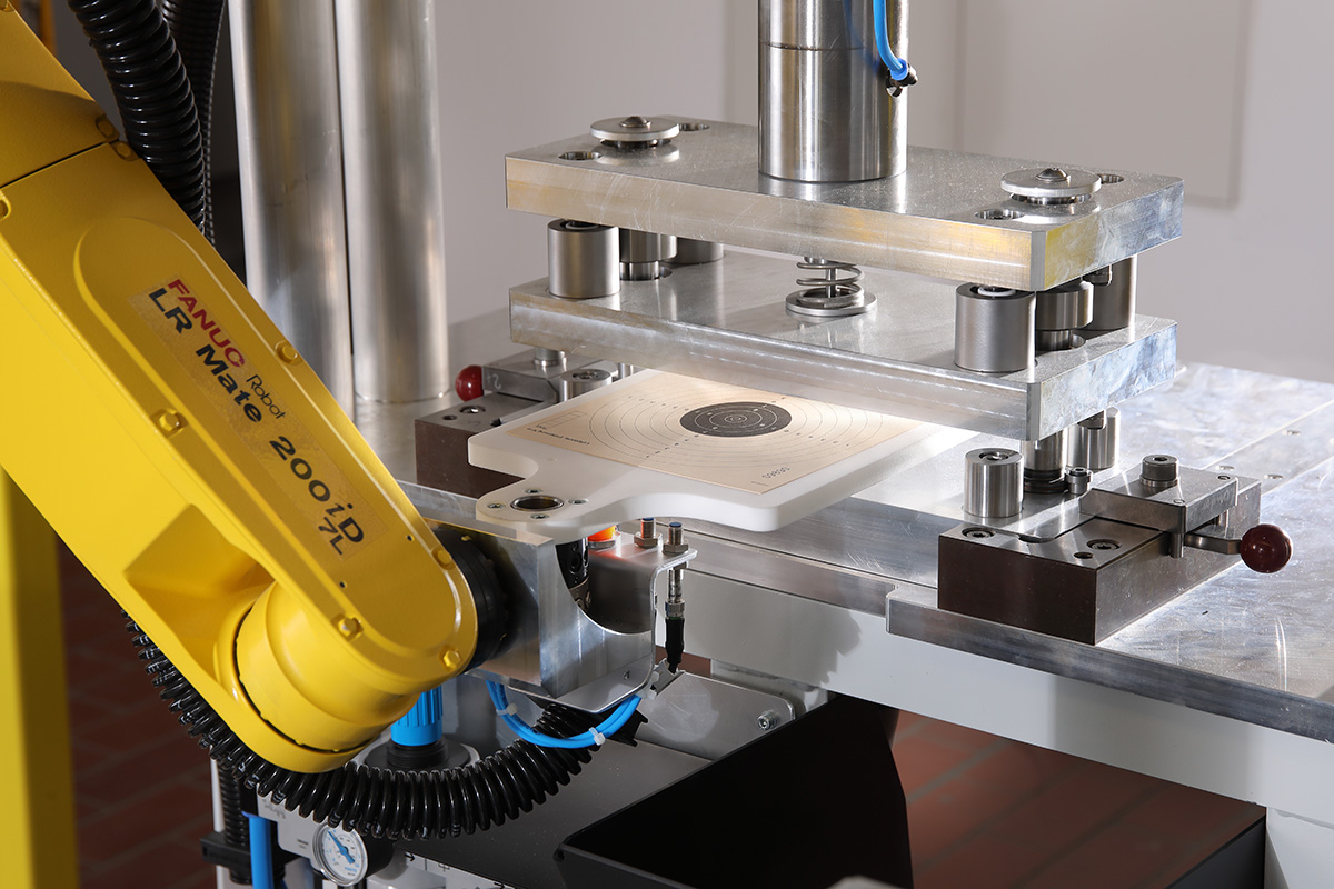 Gelber Industrieroboterarm vom Typ FANUC LR Mate 200iD/7L bedient eine Metallpresse mit einer Kreisschießscheibe aus Papier.