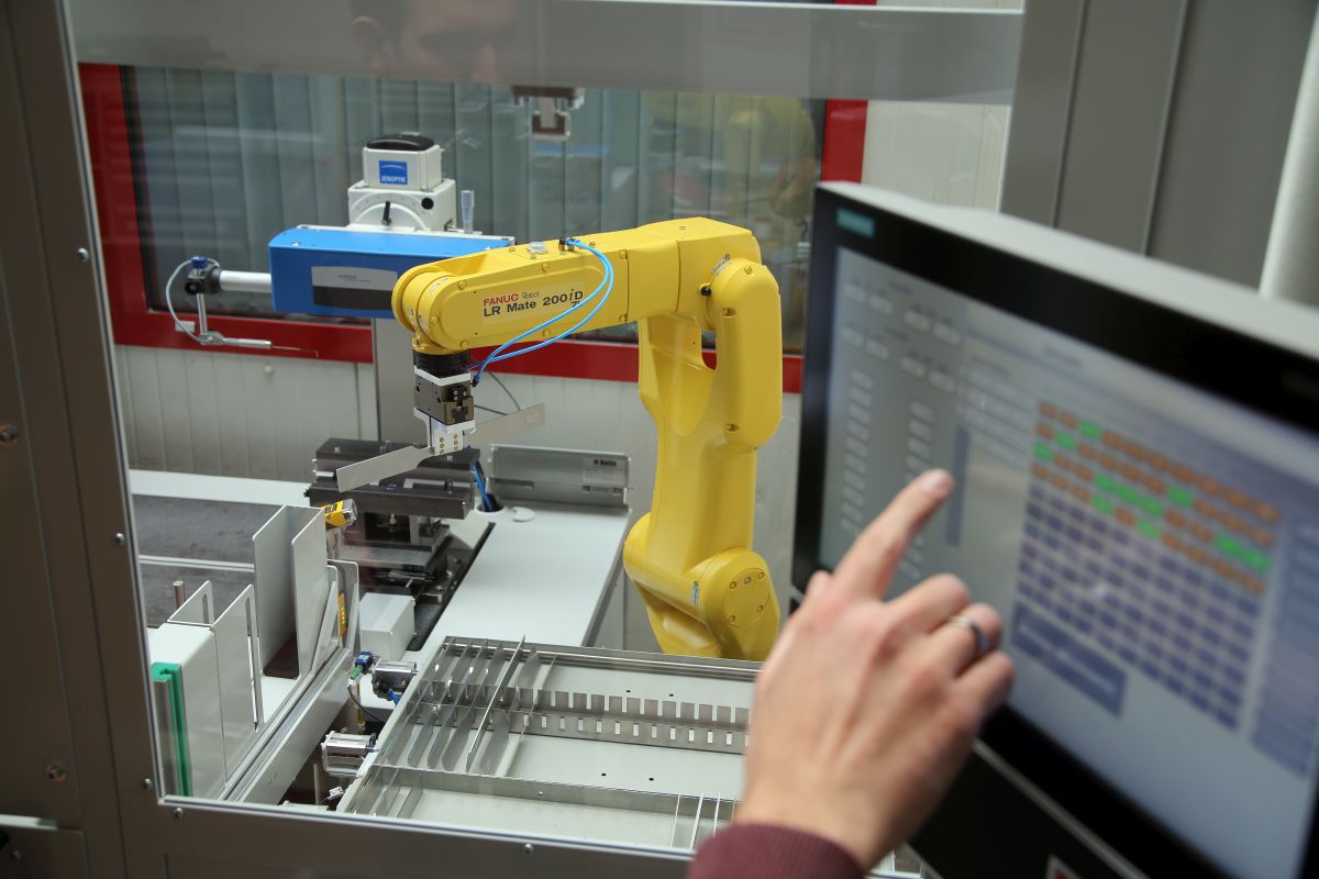 Gelber Industrieroboterarm vom Typ FANUC LR Mate 200iD in Schutzumhausung, im Vordergrund Hand an Touchscreen-Steuerung.
